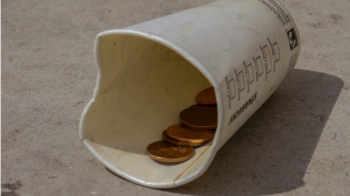 Image of a to-go coffee cup lying on a sidewalk. The inside is filled with coins and loose change. It metaphorically represents the title of the article.