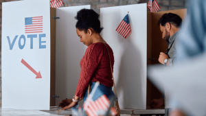 The image shows people voting in booths decorated with American flags, with a sign that says "VOTE" and an arrow pointing towards the booths