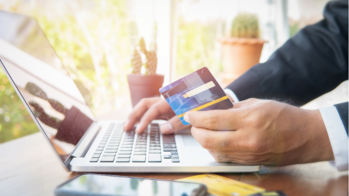 This image shows a person in a suit holding a credit card while using a laptop. A smartphone and another credit card are visible on the table.