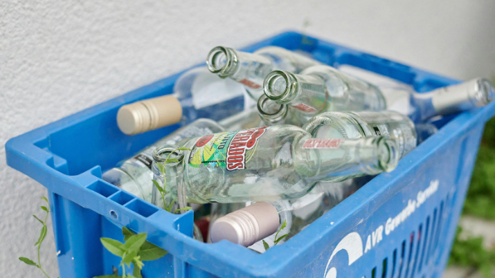 A blue recycling crate filled with empty glass bottles, including beer and liquor bottles, placed against a white wall with some green plants growing nearby